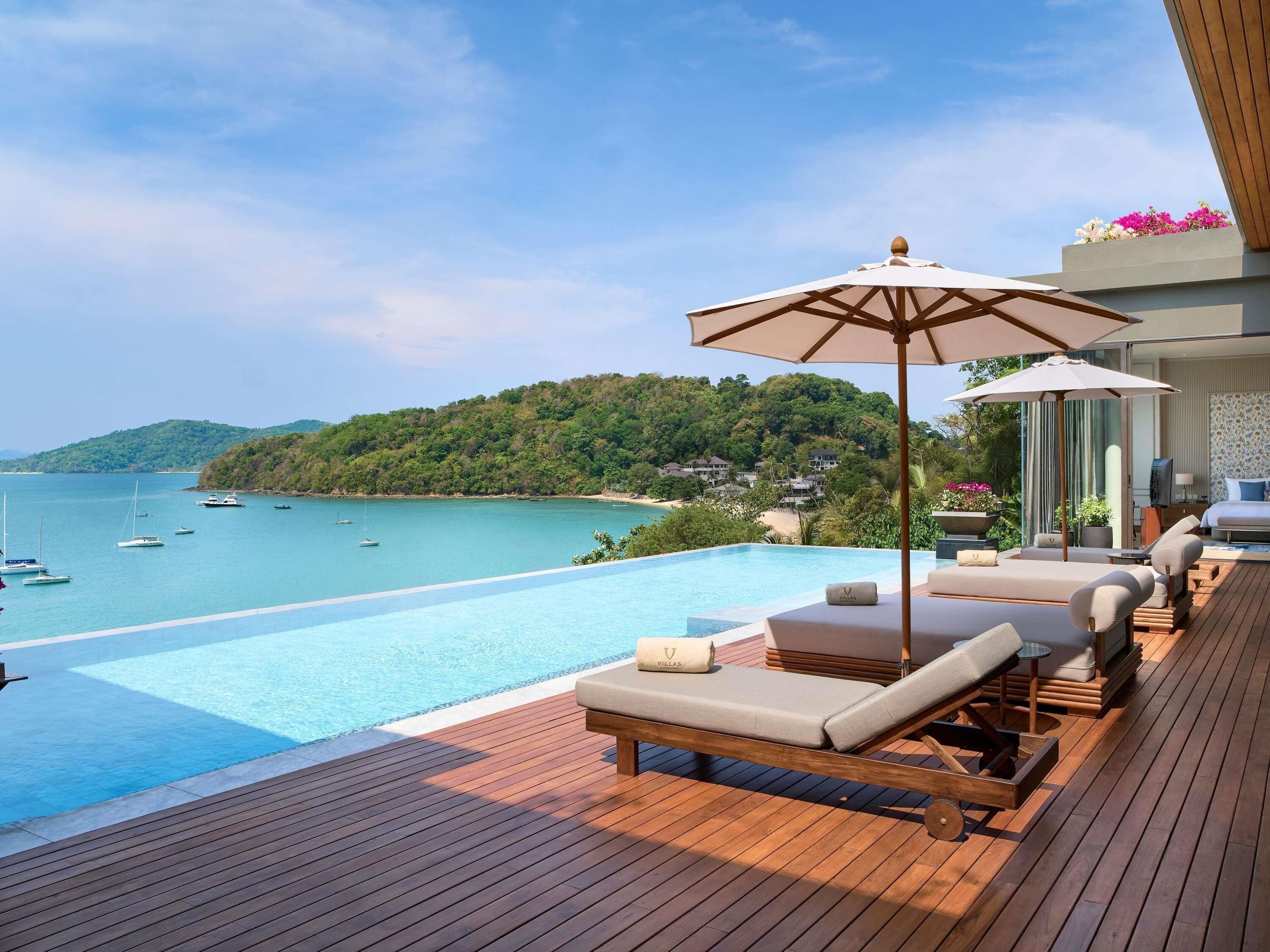 A wooden deck with a pool, lounge chairs, and a view of the sea and hills.