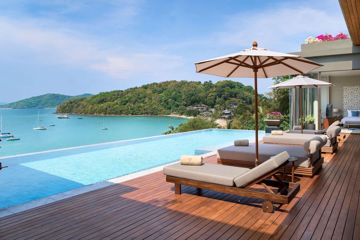A wooden deck with a pool, lounge chairs, and a view of the sea and hills.