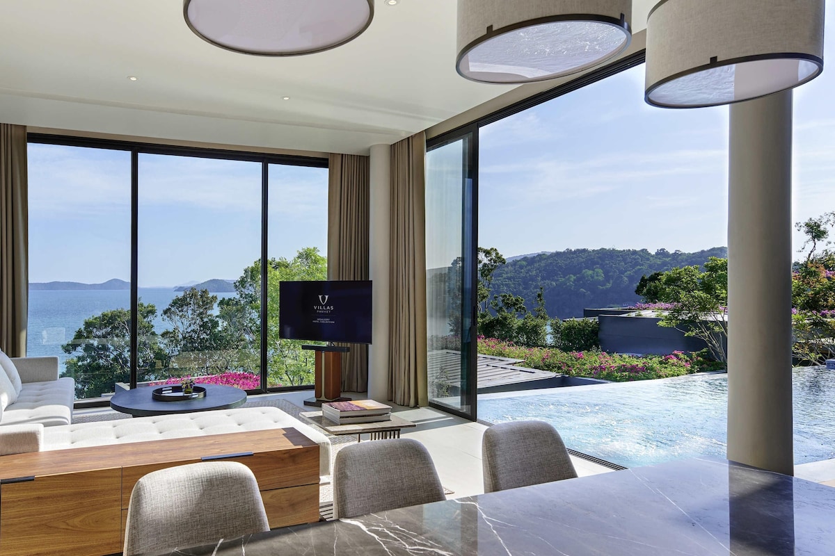 Ein modernes Hotelzimmer mit großem Fenster und Meerblick, einem Flachbildfernseher, einem Essbereich mit Marmortisch sowie Blick auf den Pool.