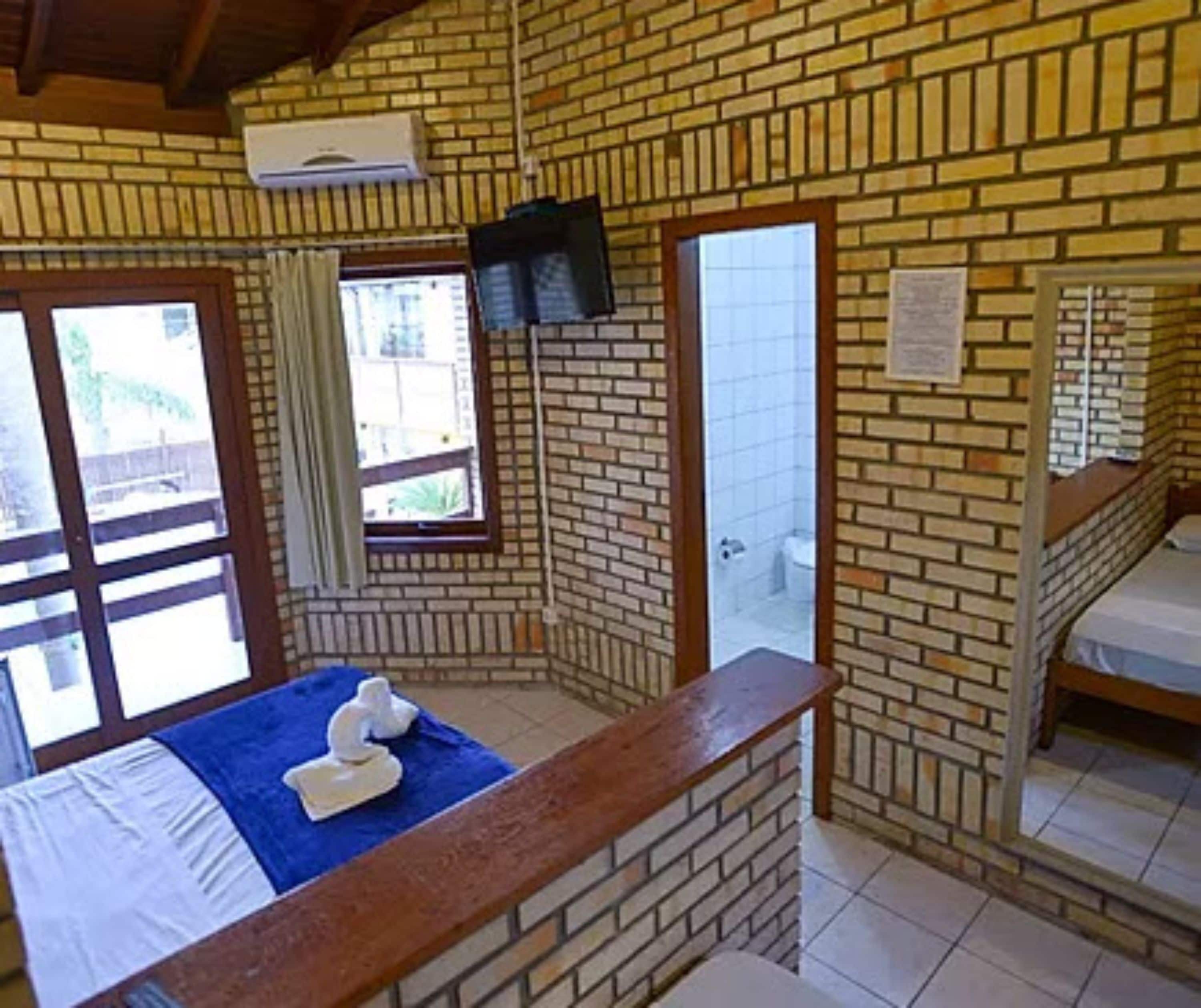 A room with a brick wall, a bed with a blue bedspread, a wooden bench, a TV mounted on the wall, and a window with curtains.
