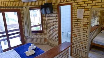 A room with a brick wall, a bed with a blue bedspread, a wooden bench, a TV mounted on the wall, and a window with curtains.