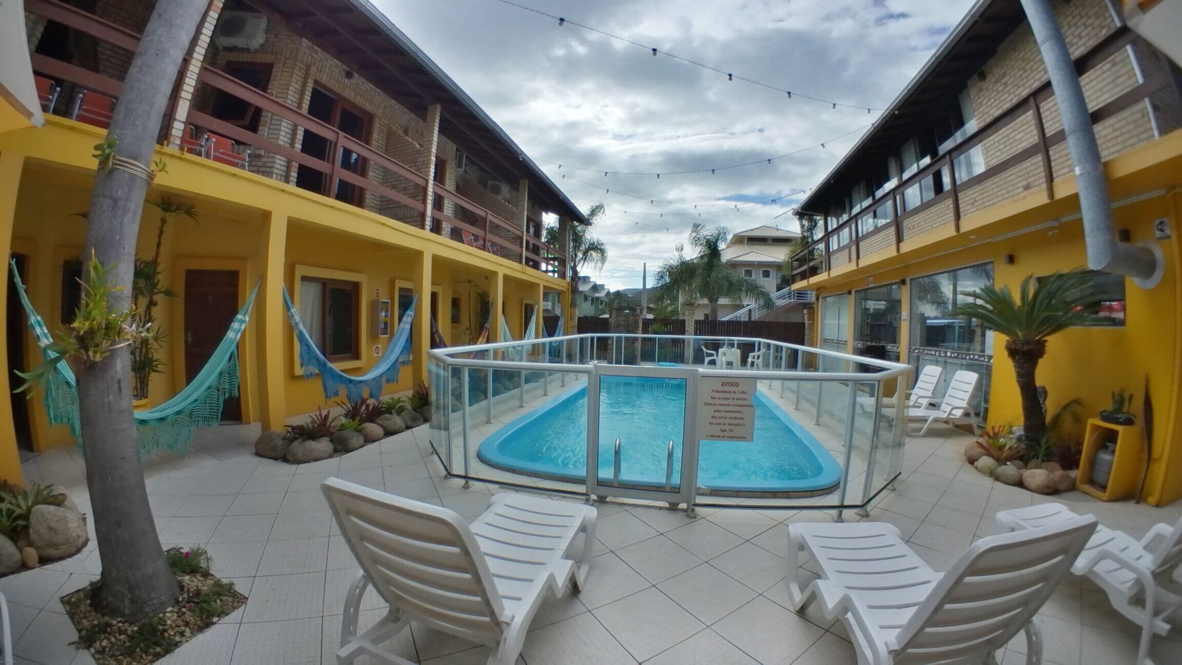 Un área de piscina con cerramiento de vidrio, rodeada de edificios amarillos, sillas de descanso blancas y una hamaca.