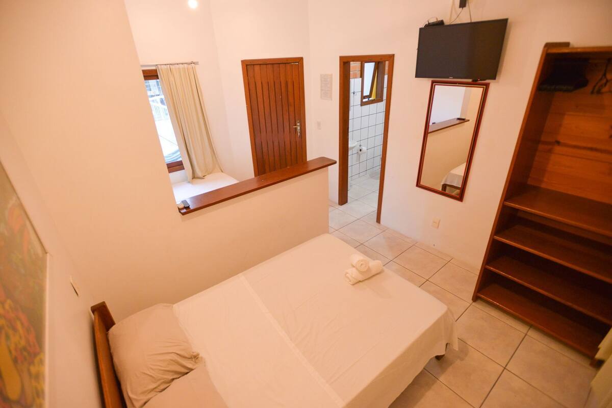 A bedroom with a bed, a window with curtains, a wooden door, a television, and a mirror.