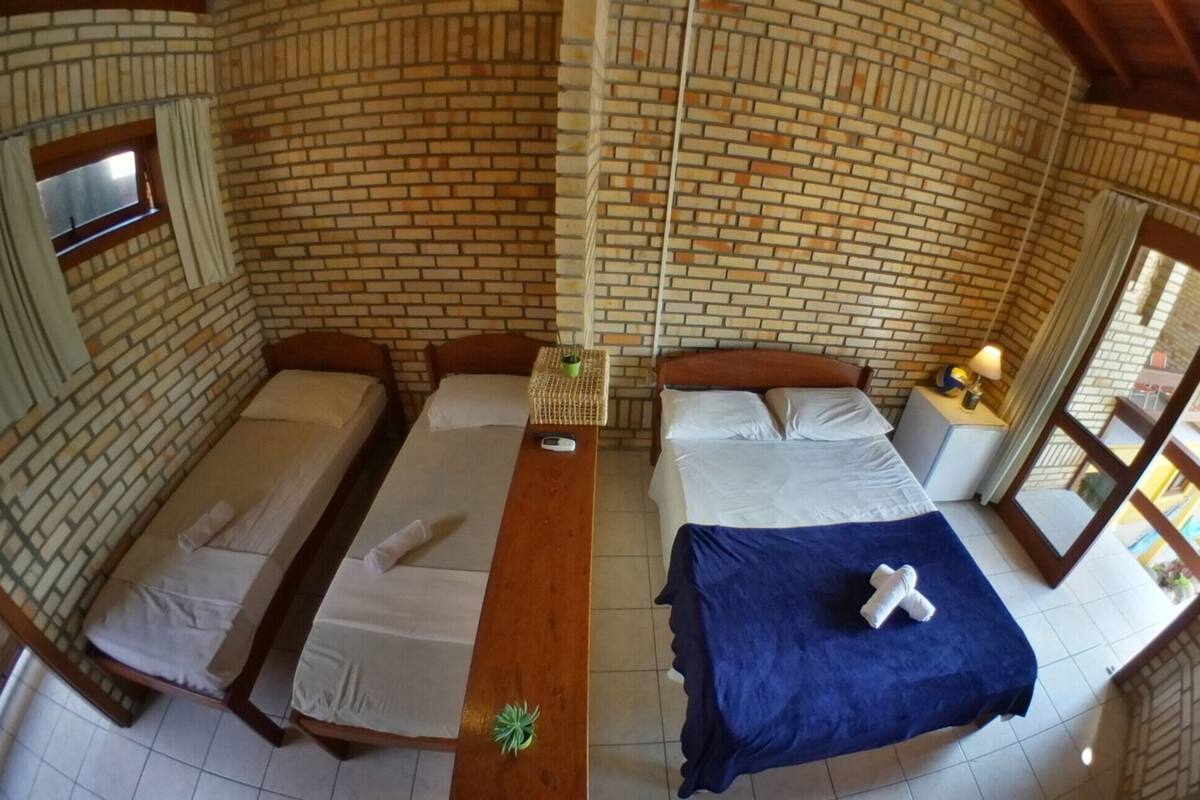 A room with a brick wall, a bed, two single beds, a wooden table, and a window.