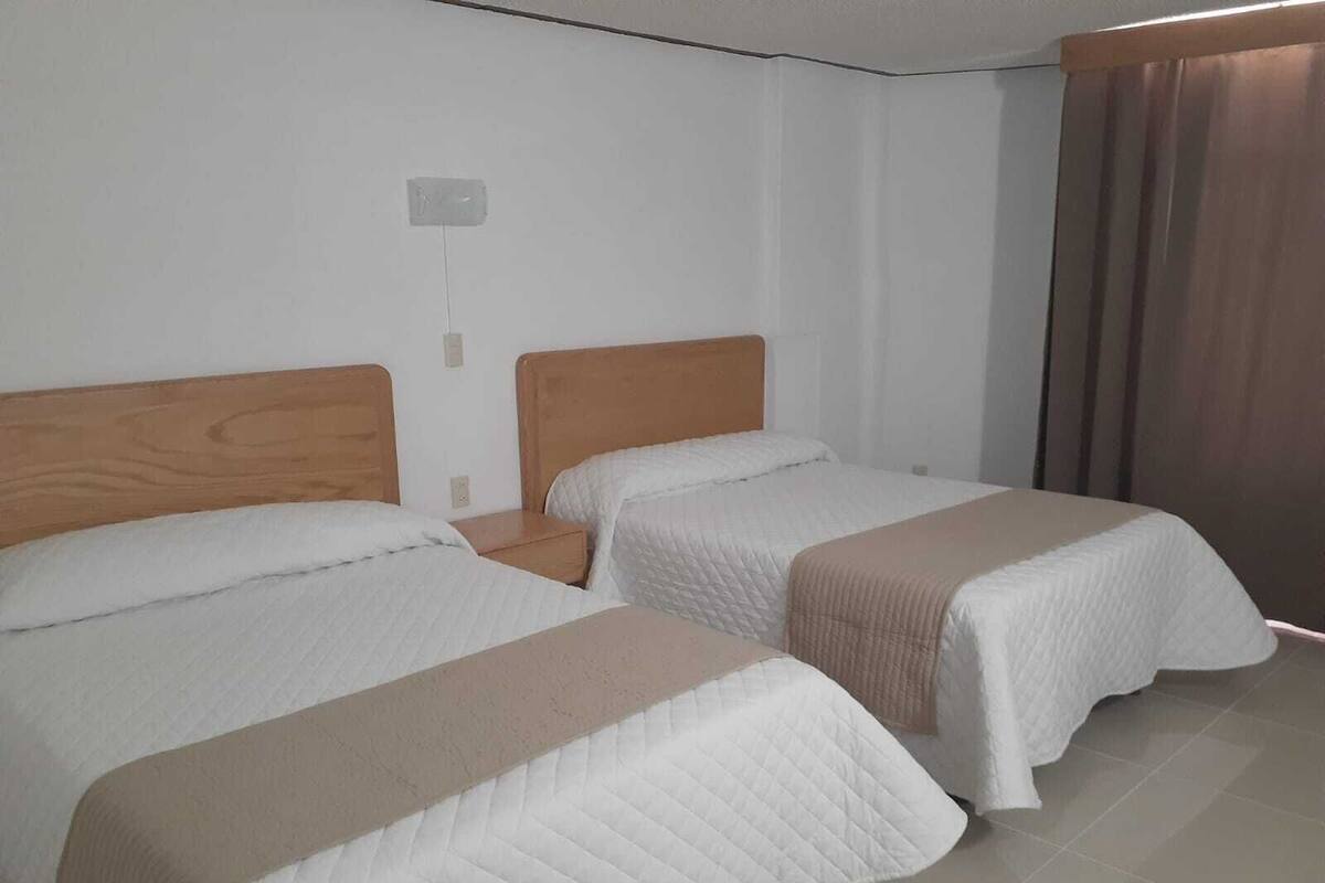 Ein Hotelzimmer mit zwei Betten, Holz-Kopfteilen, weißer Bettwäsche, einem Nachttisch und einem Fenster mit Vorhängen.