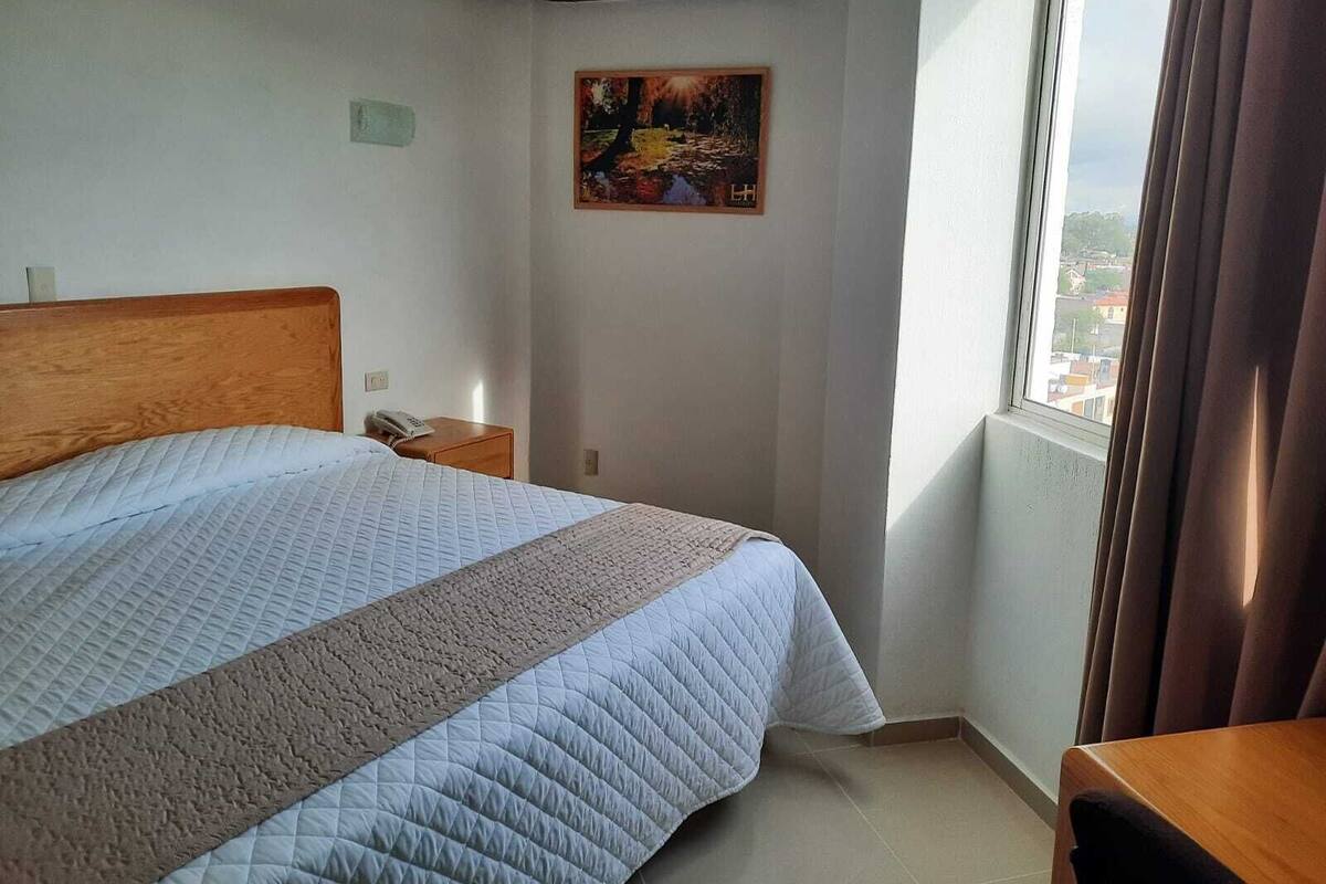 Ein Hotelzimmer mit Bett, Nachttisch, einem Bild an der Wand und einem Fenster mit Vorhängen.
