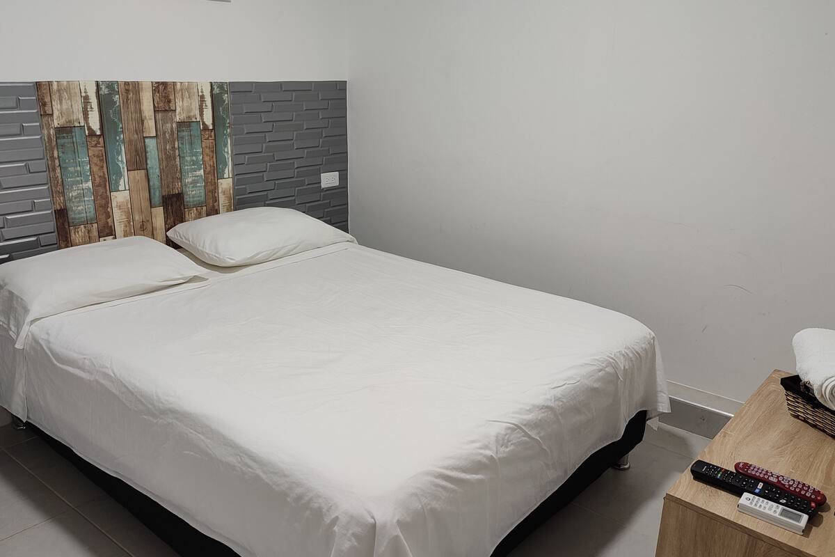 A neatly made bed with white linens, a wooden nightstand with a remote control, a rolled towel, and a potted plant.