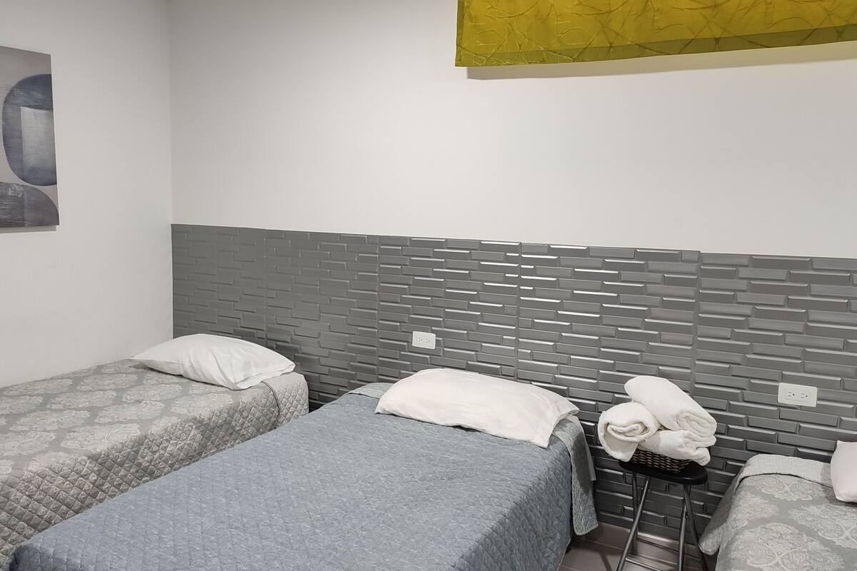 A hotel room with two beds, a grey patterned bedspread, a grey quilted blanket, a grey brick-patterned headboard, and a wall-mounted abstract painting.
