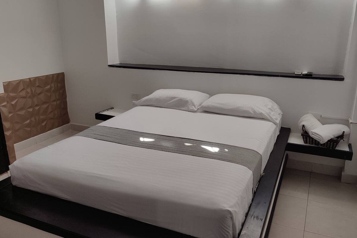 A modern hotel room with a large bed, a bedside table, a nightstand, and a wall-mounted shelf.