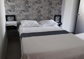 A neatly made bed with white linens, a patterned wall, and a door to the right. - Apartamentos Las Piloneras (Valledupar)