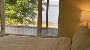 A bedroom with a large bed, a window with a view of a lake and trees, and a bedside lamp. - Fox Run Ocean Cottage (Trenton)