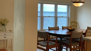 A dining area with a large window overlooking a body of water, a chandelier, and a wooden table with chairs. - Fox Run Ocean Cottage (Trenton)