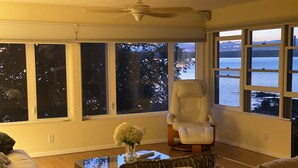 A living room with a large window overlooking a body of water, a white armchair, and a coffee table with a vase of flowers. - Fox Run Ocean Cottage (Trenton)