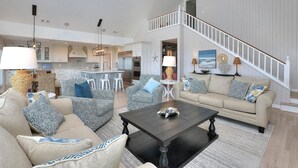 A spacious living room with a high ceiling, a staircase, and a kitchen in the background. - "Love At First Sight" - Oceanfront Home w/ Private Pool, 7 Bedrooms / 5.5 Bathrooms (Holden Beach)
