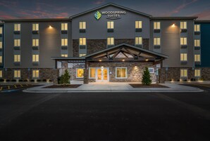 Un hotel de varios pisos con un llamativo letrero de 'Woodspring Suites', con fachada de piedra y una entrada cubierta con techo a dos aguas.