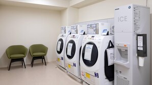 A row of four front-loading washing machines with a water dispenser and a refrigerator labeled "ICE" in a clean, modern laundry room with two green chairs.