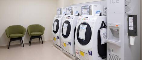 Une rangée de quatre machines à laver à chargement frontal, dotées d’un distributeur d’eau et d’un réfrigérateur portant l’inscription « GLACE », se trouve dans une buanderie propre et moderne, où sont également disposées deux chaises vertes.