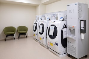 Fila de quatre rentamàquines amb càrrega frontal, un dispensador d'aigua i un refrigerador amb la inscripció "ICE" a una bugaderia moderna i immaculada amb dues cadires verdes.