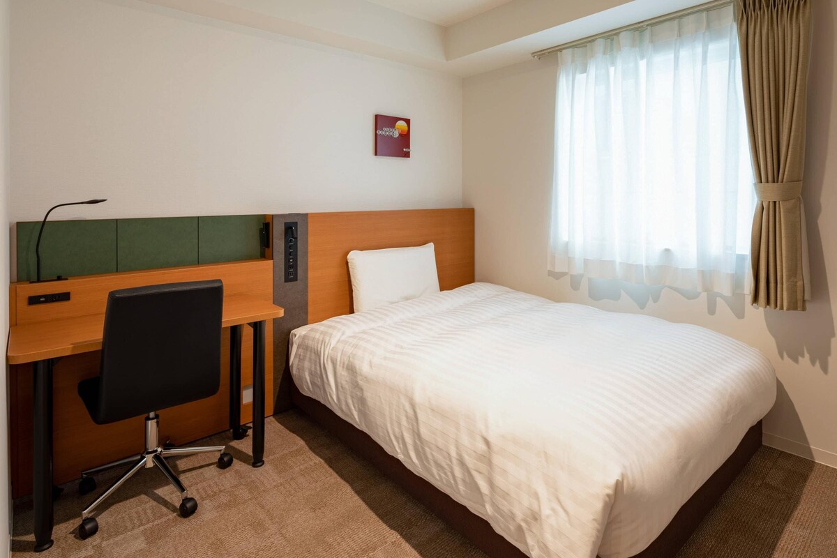 Una habitación de hotel con cama, un escritorio y una silla, y una ventana con cortinas.