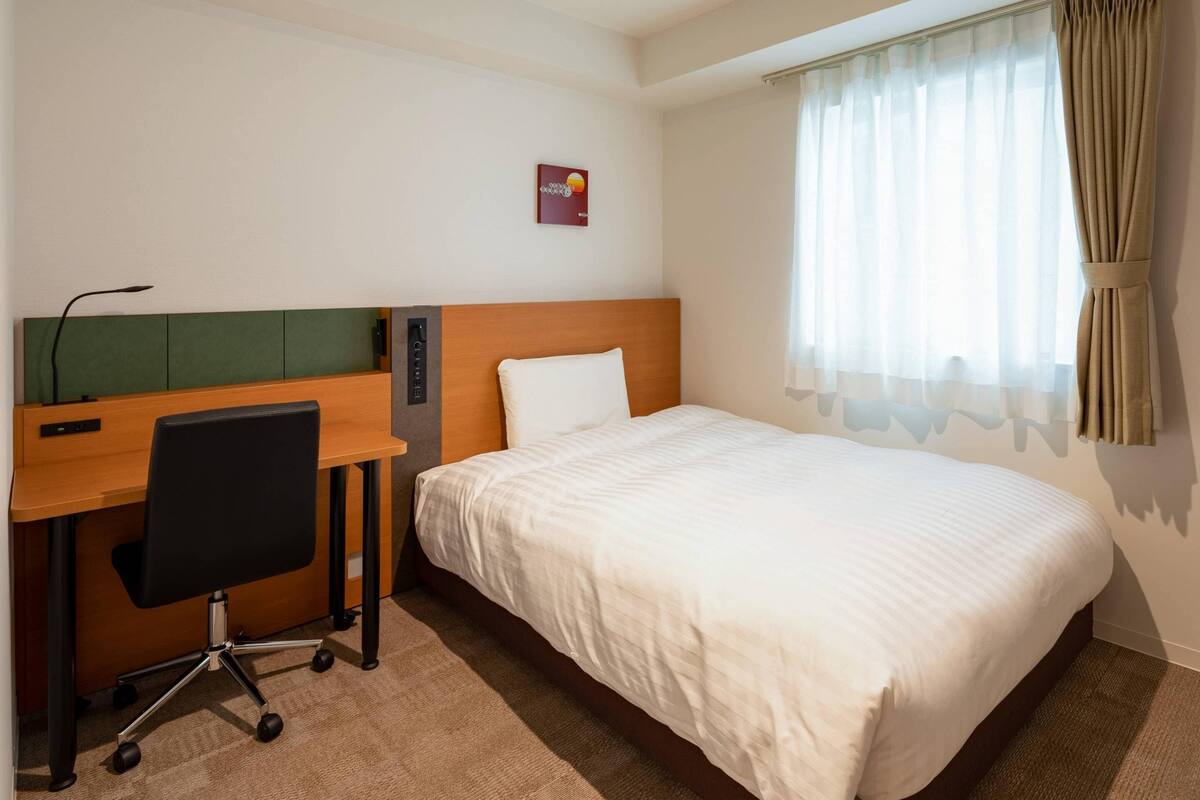 Una habitación de hotel con cama, un escritorio y una silla, y una ventana con cortinas.