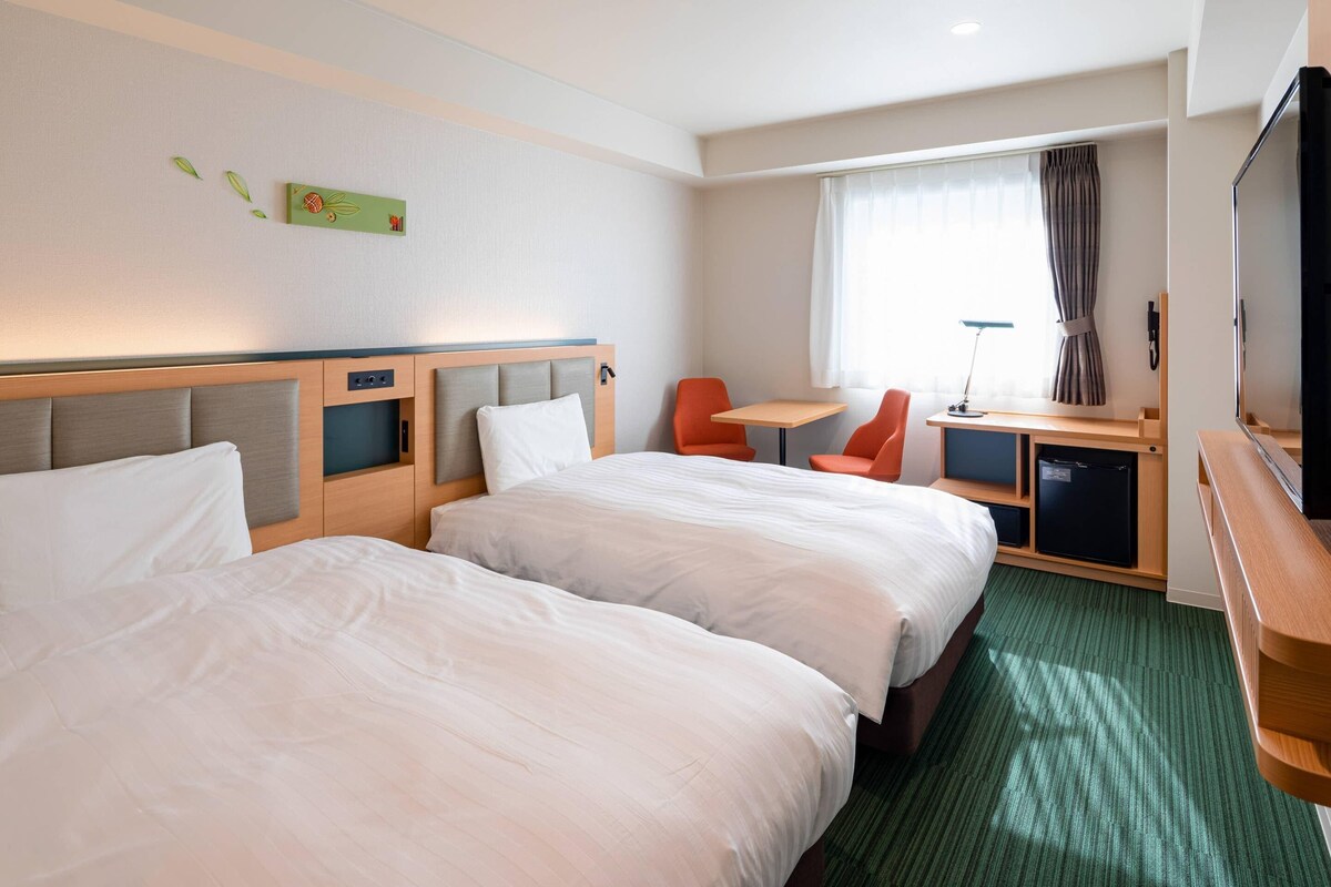 Habitación de hotel con dos camas, un escritorio, una silla, un televisor y una ventana con cortinas.