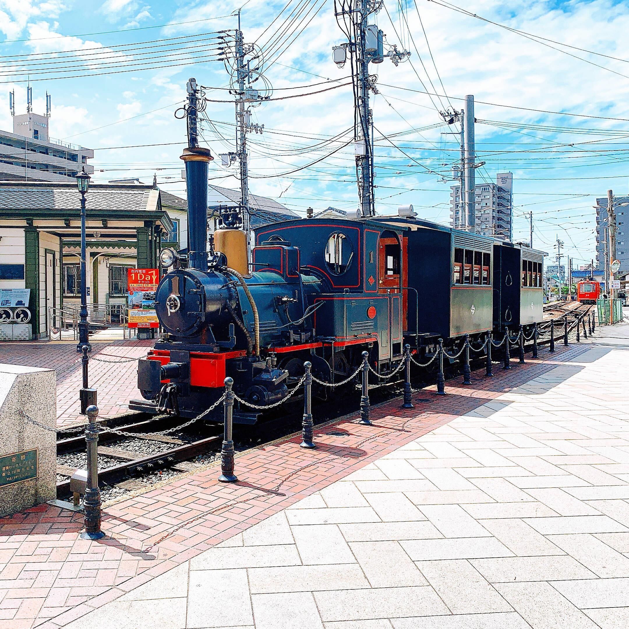 A vintage steam locomotive on display, surrounded by a chain barrier, with overhead electrical wires and a clear sky in the background.