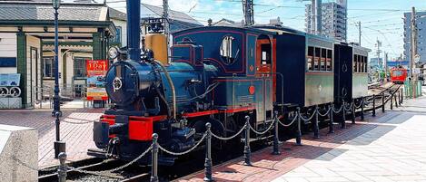 A vintage steam locomotive on display, surrounded by a chain barrier, with overhead electrical wires and a clear sky in the background.