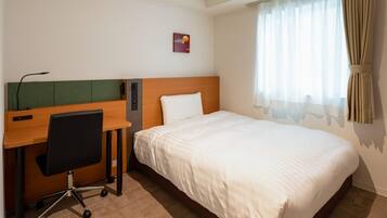 A hotel room with a bed, a desk with a chair, and a window with curtains.