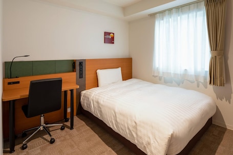 A hotel room with a bed, a desk with a chair, and a window with curtains.