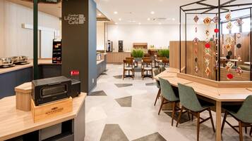 A modern library cafe with a central seating area, a kitchenette, and a decorative wall with hanging ornaments.