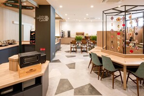 Un café moderne situé dans une bibliothèque, avec un espace de repos central, un coin cuisine et un mur décoratif orné d’objets suspendus.