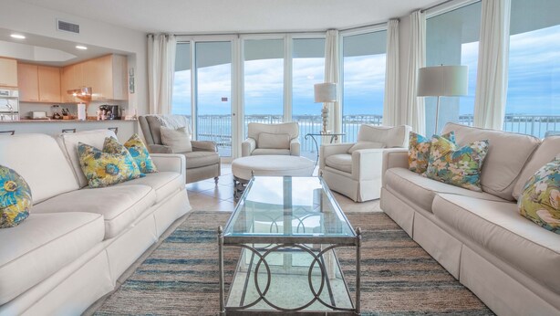 Ein geräumiges Wohnzimmer mit einem Glastisch, zwei Sofas mit floralen Kissen, einem runden Hocker und großen Fenstern mit Meerblick.
