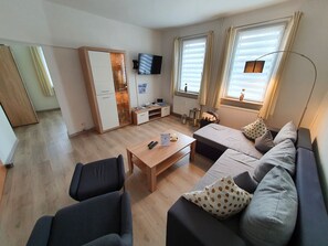 En moderne stue med sofa, to lænestole, sofabord og tv på væggen. Der er et træskab med glaslåger, et lille bord med lampe og et vindue med gardiner.