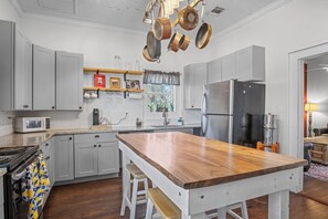 A kitchen with a central island, grey cabinets, a stainless steel refrigerator, and hanging pots. - Mighty Oaks Retreat | 2BR Farmhouse w/ Fire Pit (D'Hanis)