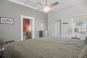 Ein Schlafzimmer mit einem Bett, einem Deckenventilator, einer Tür, einem Fenster mit Jalousien und einer Wand mit gerahmten Bildern.