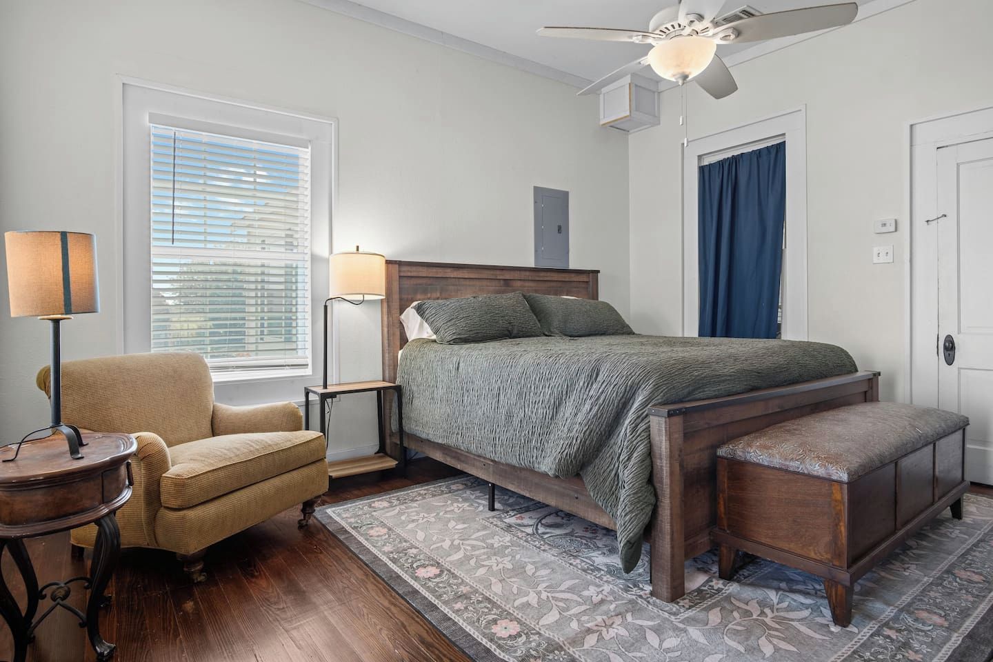 A bedroom with a bed, a chair, a nightstand, a lamp, a ceiling fan, and a window with blinds.