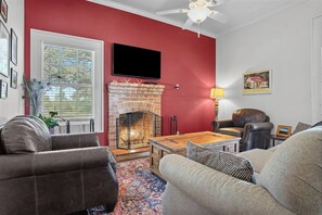 A living room with a fireplace, leather sofas, a television, and a ceiling fan. - Mighty Oaks Retreat | 2BR Farmhouse w/ Fire Pit (D'Hanis)