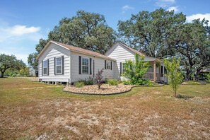 A two-story house with a front porch and a small garden area. - Mighty Oaks Retreat | 2BR Farmhouse w/ Fire Pit (D'Hanis)