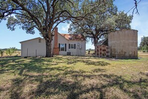 A single-story house with a brick chimney, a large tree in front, and a rusted metal structure to the right. - Mighty Oaks Retreat | 2BR Farmhouse w/ Fire Pit (D'Hanis)