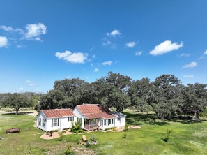 A two-story house with a porch, surrounded by a grassy yard and trees. - Mighty Oaks Retreat | 2BR Farmhouse w/ Fire Pit (D'Hanis)