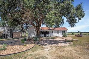 A house with a large tree in front, a gravel driveway, and a metal barrel in the yard. - Mighty Oaks Retreat | 2BR Farmhouse w/ Fire Pit (D'Hanis)