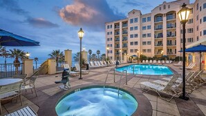 Zona de piscina hotelera con sillones de descanso, una bañera de hidromasaje y un edificio grande al fondo.