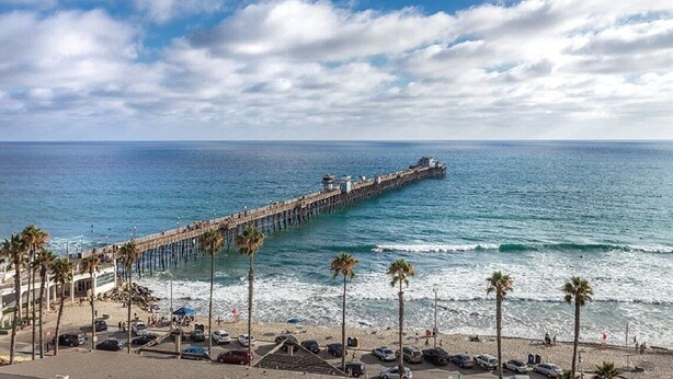 Un muelle que se adentra en el océano, con una playa y coches aparcados al frente.