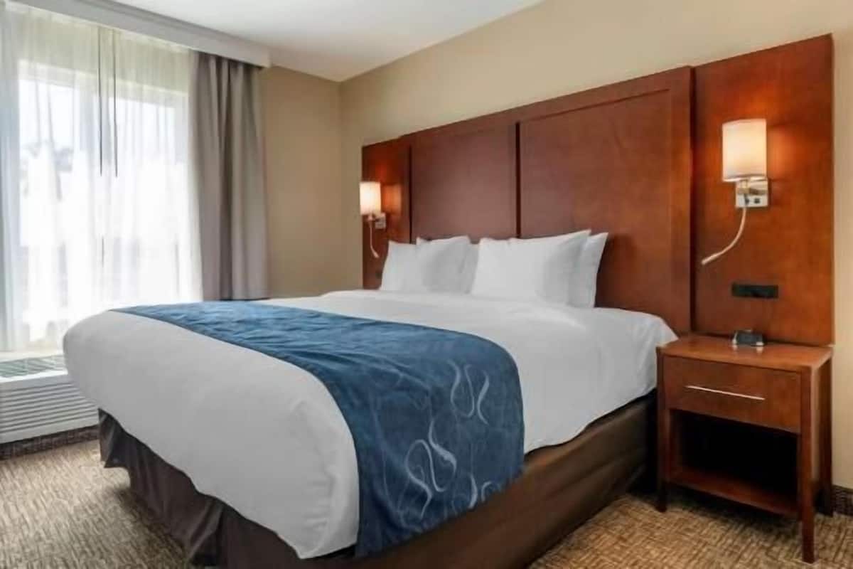 A hotel room with a large bed, a wooden headboard, two wall-mounted lamps, a nightstand, and a window with curtains.