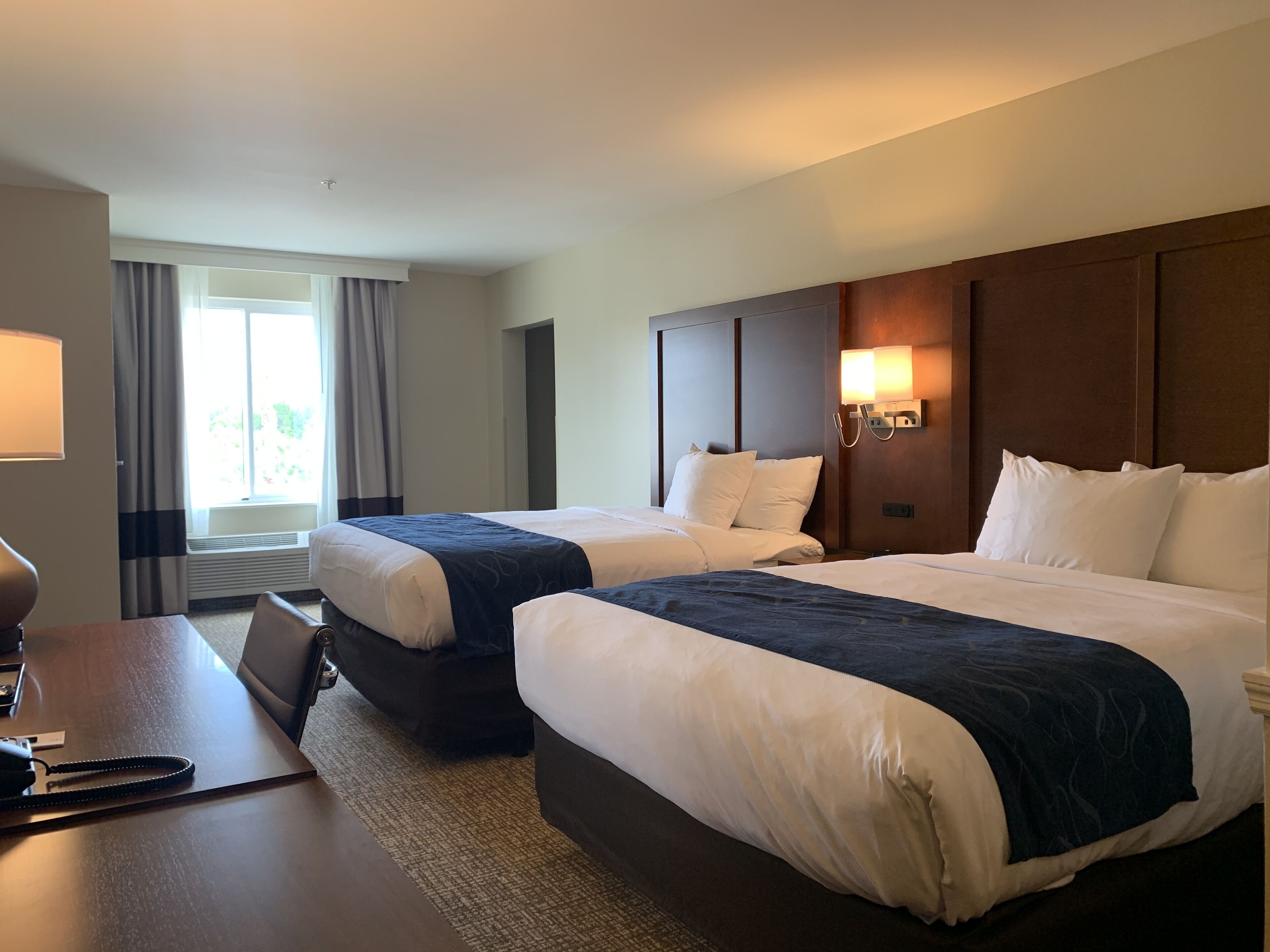 Ein Hotelzimmer mit zwei Betten, einem Schreibtisch, einer Lampe, einem Fenster mit Vorhängen und einer Wandleuchte.