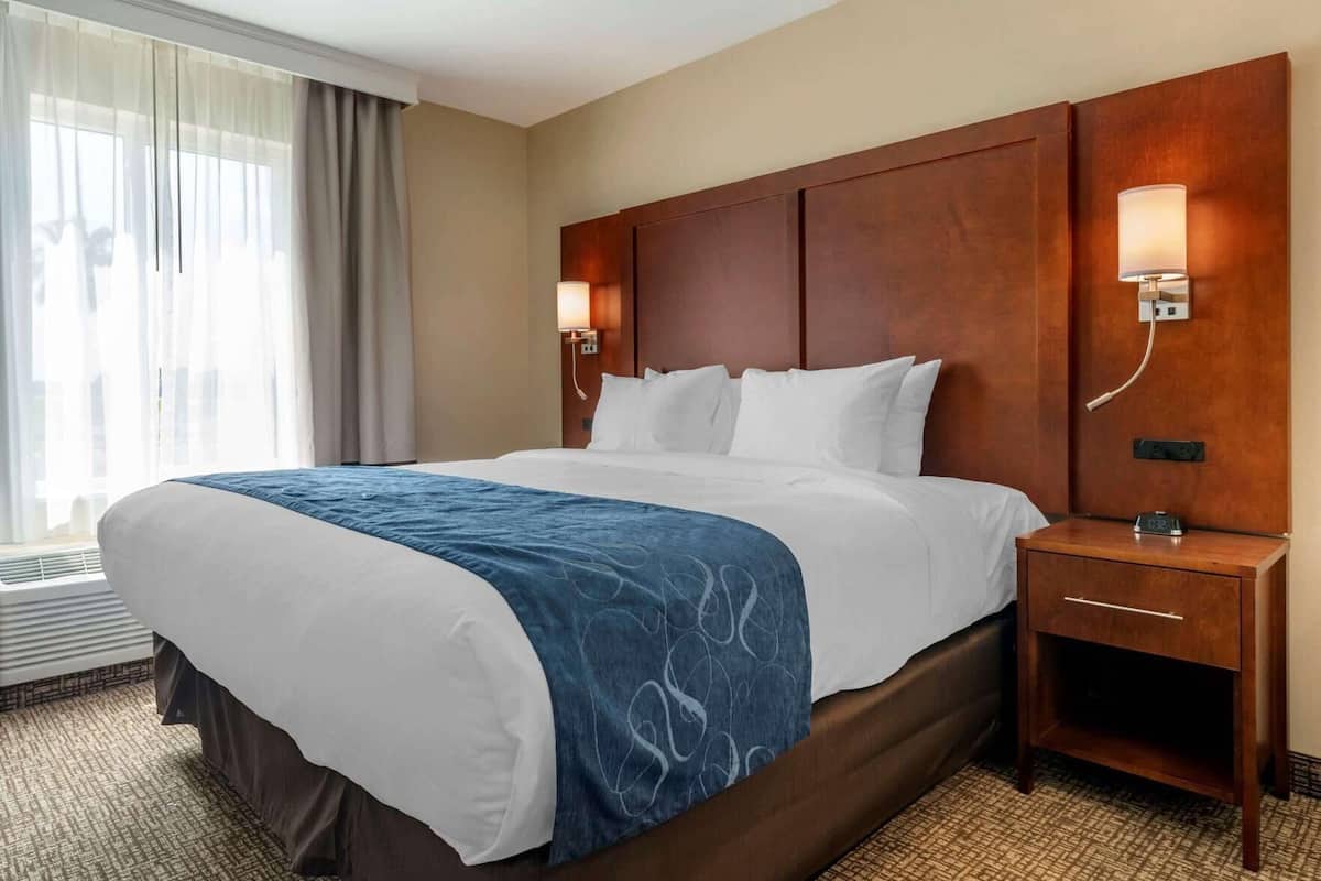 A hotel room with a large bed, a wooden headboard, two bedside lamps, a nightstand, and a window with curtains.