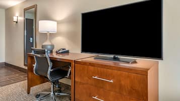A hotel room with a wooden desk, a black office chair, a flat-screen TV, a lamp, and a mirror.