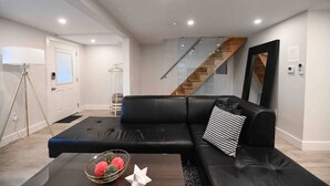 Una sala de estar moderna con un sofá de cuero negro, una mesa de centro de vidrio con objetos decorativos, una lámpara de pie y una escalera que conduce a otro nivel.