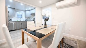Una cocina moderna con electrodomésticos de acero inoxidable, un comedor con mesa de vidrio y sillas blancas, y un pequeño jarrón con flores sobre la mesa.