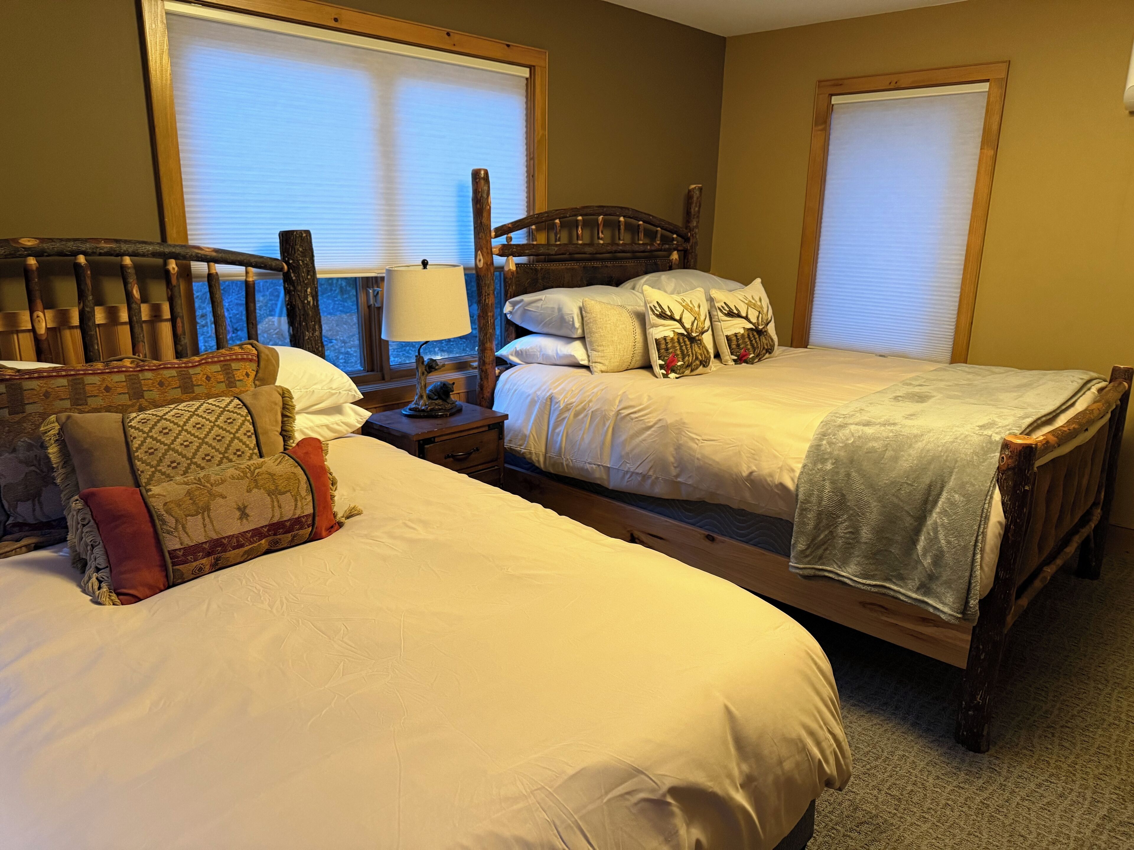 A bedroom with two beds, a nightstand with a lamp, and a window with blinds.